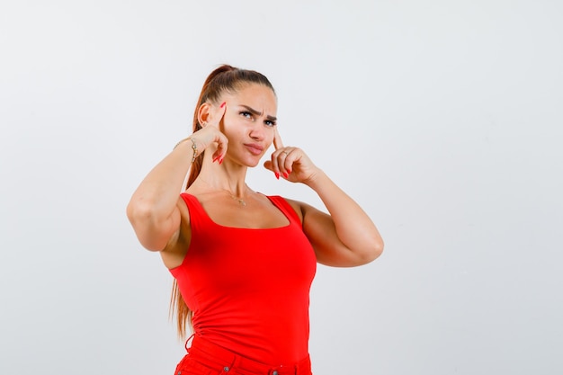 Hembra joven sosteniendo los dedos en las sienes en camiseta sin mangas roja y mirando pensativo, vista frontal.