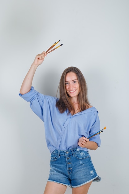 Hembra joven sosteniendo suministros de pintura en camisa, pantalones cortos y mirando alegre