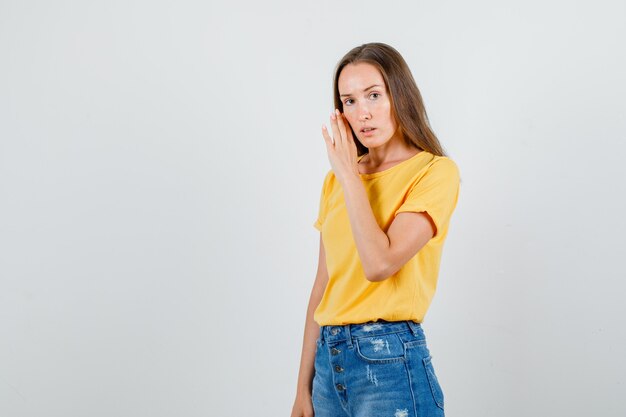 Hembra joven susurrando secreto detrás de la mano en camiseta, pantalones cortos y mirando serio