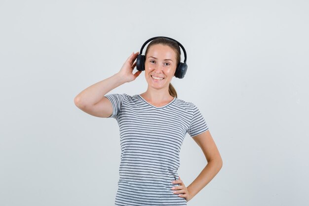 Hembra joven tocando auriculares en camiseta a rayas y mirando alegre, vista frontal.