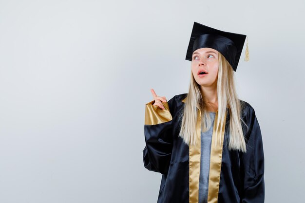 Hembra joven en uniforme graduado apuntando hacia arriba y mirando enfocado