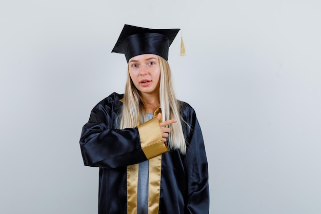 Hembra joven en uniforme graduado apuntando a un lado y mirando asombrado