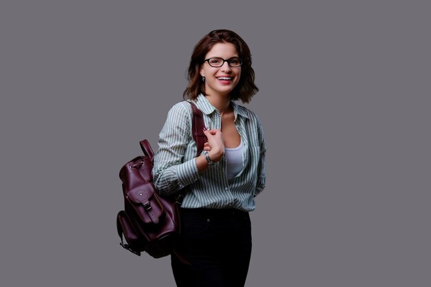 Hembra joven viajera positiva con mochila de cuero aislada sobre fondo gris.