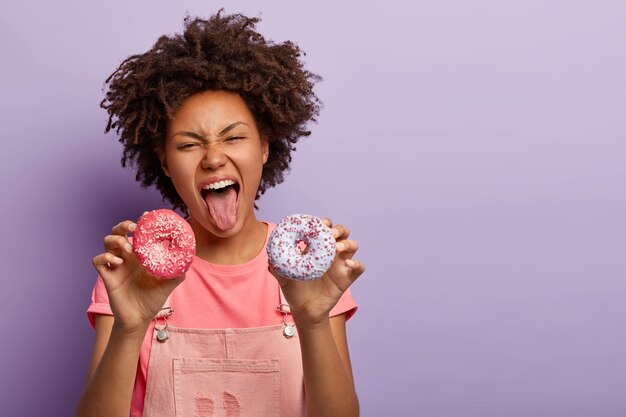 La hembra juguetona de piel oscura mantiene la boca abierta y muestra la lengua, tiene pasión por los deliciosos donuts glaseados, rompe la dieta y tiene una nutrición poco saludable, vestida de manera informal, disfruta de un delicioso refrigerio. Dulce vida