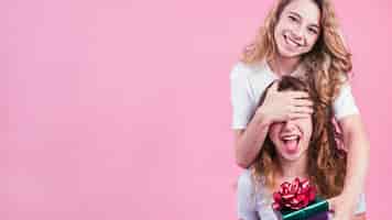 Foto gratuita hembra que cubre los ojos de su amigo que da la caja de regalo contra fondo rosado