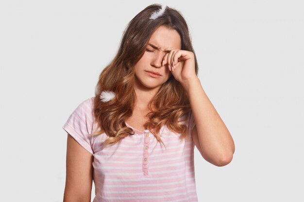 la hembra soñolienta frota los ojos, odia levantarse temprano, tiene plumas en la cabeza, vestida con pijama, aislada en blanco. Joven insomne trabajó mucho durante la noche. Hora de la mañana