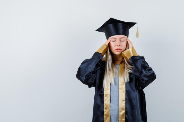 Las hembras jóvenes frotándose las sienes en uniforme de posgrado y con aspecto cansado