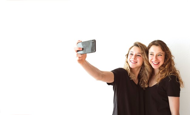 Hermana sonriente que toma el selfie en el teléfono móvil contra el contexto blanco