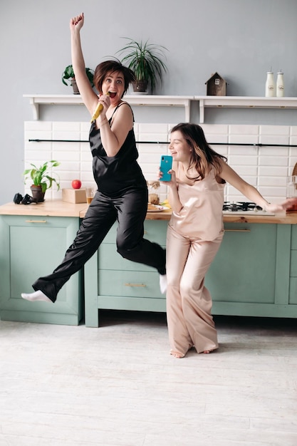 Hermanas atractivas en pijamas en blanco y negro escuchando música y bailando en la cocina por la mañana Felices chicas guapas divirtiéndose mientras cocinan el desayuno Amigos morenas cantando y saltando