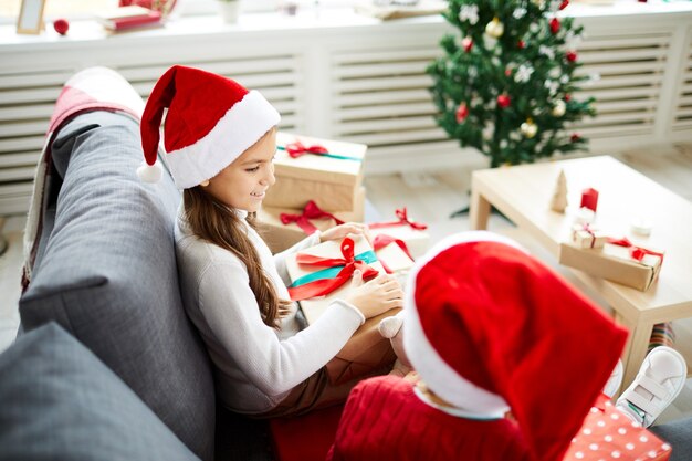 Hermanas felices sentados en el sofá y desenvolver regalos de Navidad