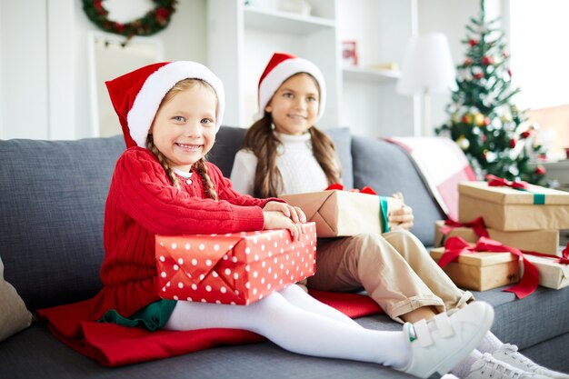 Hermanas felices sentados en el sofá y desenvolver regalos de Navidad