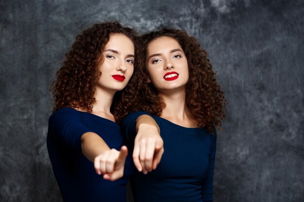 Hermanas gemelas sonriendo señalando con el dedo a la cámara en gris