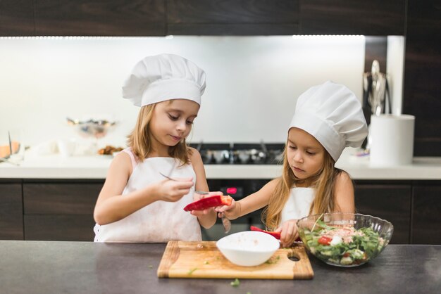 Hermanas lindas adorables en el sombrero y los delantales del cocinero que preparan la comida en cocina