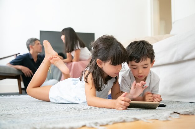 Hermano y hermana pequeños acostados en el piso de la sala de estar y usando dispositivos digitales con aplicaciones de aprendizaje mientras los padres se sientan juntos