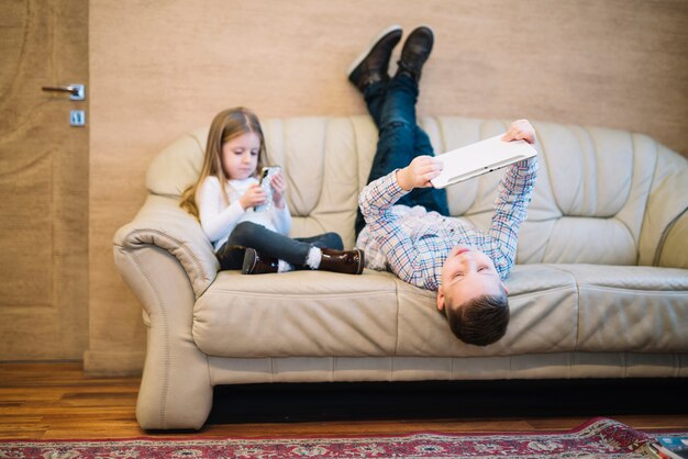 Hermano y hermana sentados en el sofá usando un teléfono móvil y una tableta digital