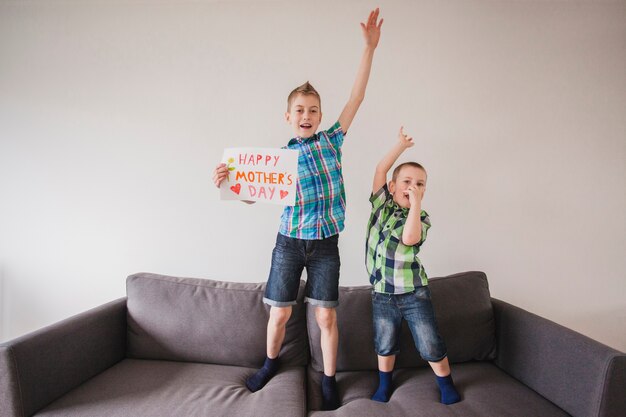 Hermanos de pié en el sofá con cartel para el día de la madre