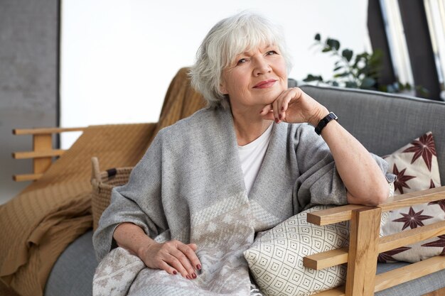 Hermosa abuela aseada de sesenta años con amplia bufanda gris y reloj de pulsera descansando cómodamente en el sofá de la sala de estar, sonriendo felizmente, esperando a que lleguen su hijo y sus nietos