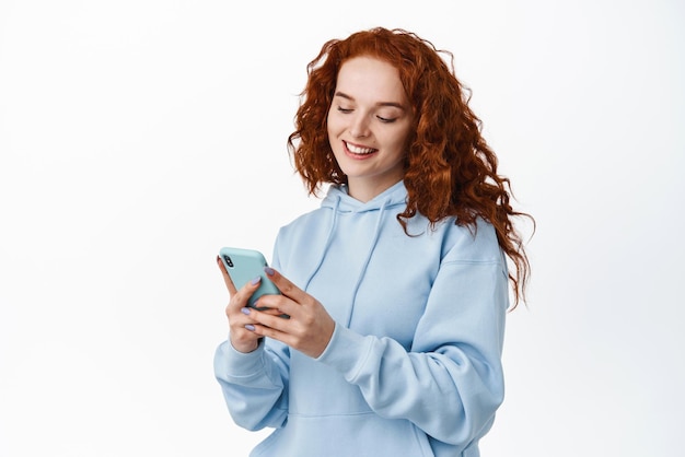 Hermosa adolescente pelirroja usando la aplicación de chat de teléfono móvil mirando el teléfono inteligente leyendo la pantalla del móvil y sonriendo de pie contra el fondo blanco