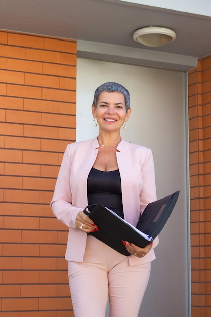 Hermosa agente inmobiliaria femenina parada en la entrada de la casa. Mujer sonriente con el pelo corto y gris en traje rosa preparándose para conocer a los clientes. Bienes raíces, negocios, concepto de trabajo.