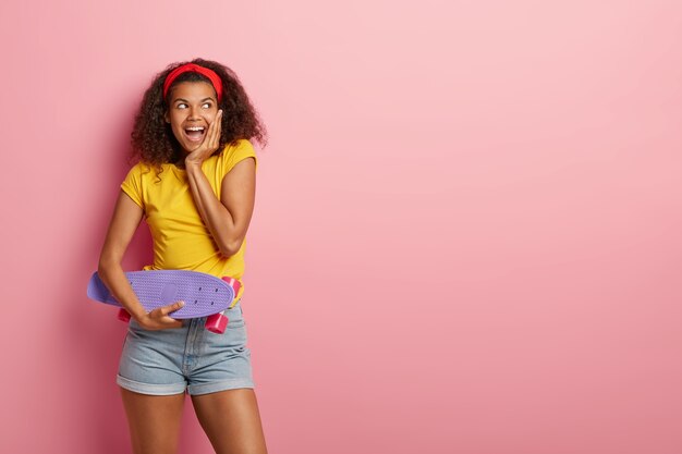 Hermosa alegre adolescente afroamericana intenta mostrar lo mejor de sí misma en el skate, sostiene el tablero de centavo púrpura