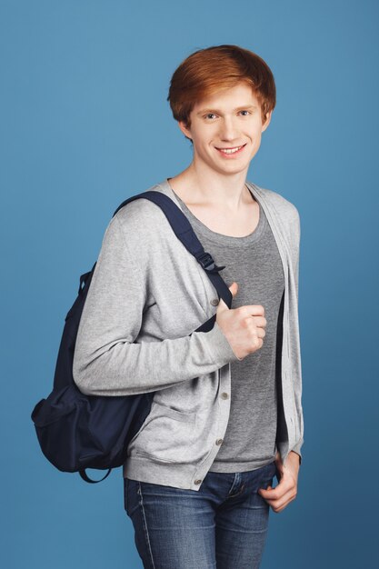 Hermosa y alegre estudiante alegre con cabello pelirrojo en ropa casual sonriendo, sosteniendo la mochila y la mano en el bolsillo, con expresión relajada y feliz.