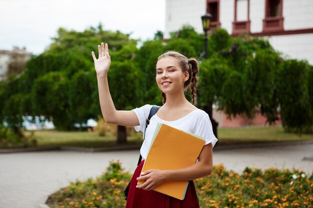 Hermosa alegre estudiante sonriendo, saludando, sosteniendo carpetas al aire libre