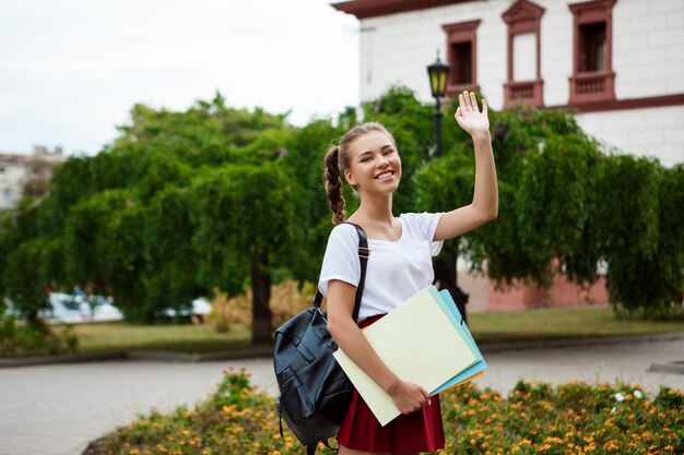 Hermosa alegre estudiante sonriendo, saludando, sosteniendo carpetas al aire libre