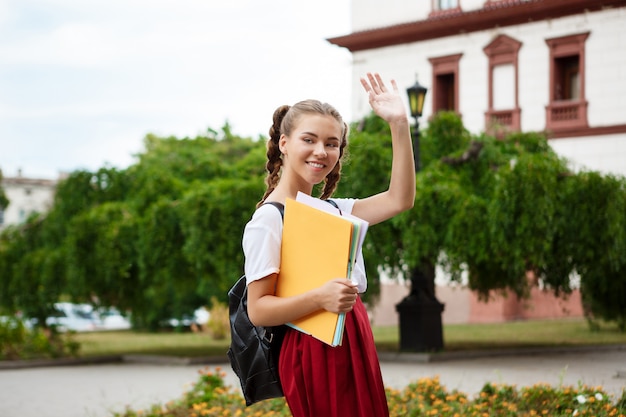 Hermosa alegre estudiante sonriendo, saludando, sosteniendo carpetas al aire libre