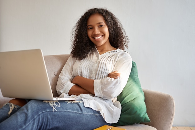 Hermosa alegre joven afroamericana confiada sentada en el sofá con la computadora portátil, cruzando los brazos sobre el pecho, viendo el video blog en línea, con una amplia sonrisa con dientes