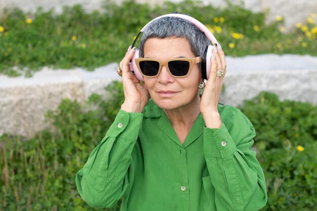 Hermosa anciana escuchando música en el parque. Modelo femenino con cabello gris corto en ropa brillante y grandes auriculares. Ocio, actividad, concepto de tecnología.