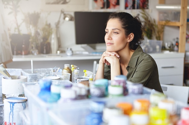 Hermosa artista femenina con expresión pensativa, sentada en su lugar de trabajo con acuarelas, tratando de imaginar la imagen que ella va a pintar. Gente, hobby, creatividad, concepto de pintura