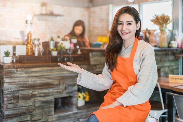 Hermosa y atractiva dueña de una cafetería asiática sonríe con felicidad y alegría con un delantal de taza de café en el fondo del café