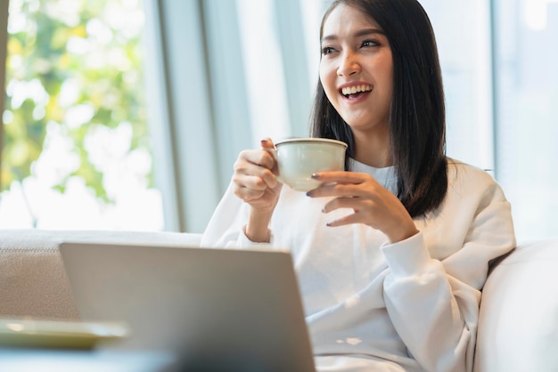 Hermosa y atractiva frescura femenina asiática sonrisa alegre mañana trabajando con café portátil desde casa con felicidad disfrutar de la vista al jardín mujer joven asiática con fondo de jardín de luz de ventana