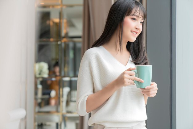 Hermosa y atractiva frescura femenina asiática sonrisa alegre mañana trabajando con café portátil desde casa con felicidad disfrutar de la vista al jardín mujer joven asiática con fondo de jardín de luz de ventana