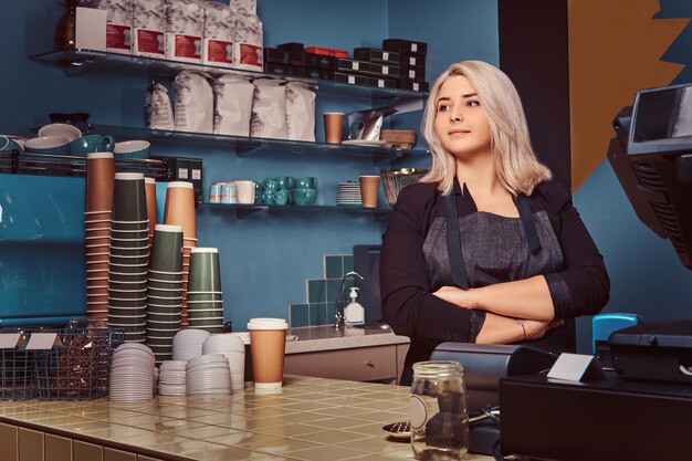 Hermosa barista experimentada en delantal de pie con los brazos cruzados en la cafetería.