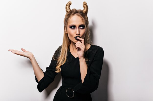 Hermosa bruja en vestido posando en el carnaval. Mujer rubia pensativa en traje de vampiro celebrando halloween.