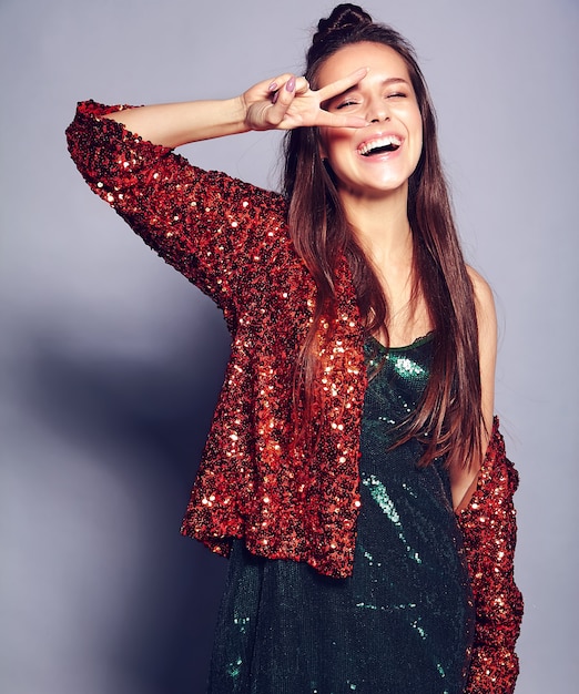 Hermosa caucásica sonriente hipster morena mujer modelo en brillante brillante reflejando verano elegante chaqueta y vestido verde posando en gris. Mostrando signo de paz