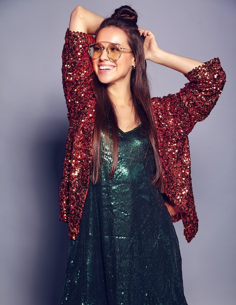 Hermosa caucásica sonriente hipster mujer morena modelo en brillante brillante que refleja la elegante chaqueta de verano y el vestido verde posando en gris