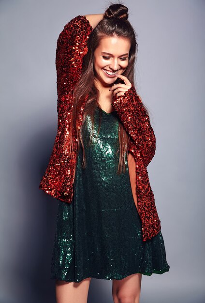 Hermosa caucásica sonriente hipster mujer morena modelo en brillante brillante que refleja la elegante chaqueta de verano y el vestido verde posando en gris