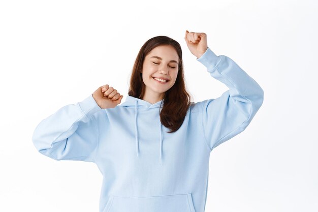 Hermosa chica adolescente bailando en su sudadera con capucha, levantando las manos y sonriendo alegre, posando y divirtiéndose contra el fondo blanco.