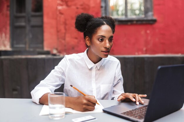 Hermosa chica africana con camisa blanca trabajando soñadoramente con una laptop en un acogedor patio de café
