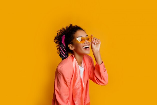 Hermosa chica africana en chaqueta rosa posando con expresión de la cara feliz sobre fondo amarillo.