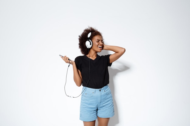 Hermosa chica africana escuchando música en auriculares sobre la pared blanca.