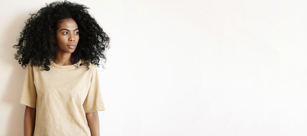Hermosa chica africana con peinado afro con camiseta de gran tamaño de pie aislado en la pared blanca y mirando a otro lado con expresión seria y pensativa en su rostro