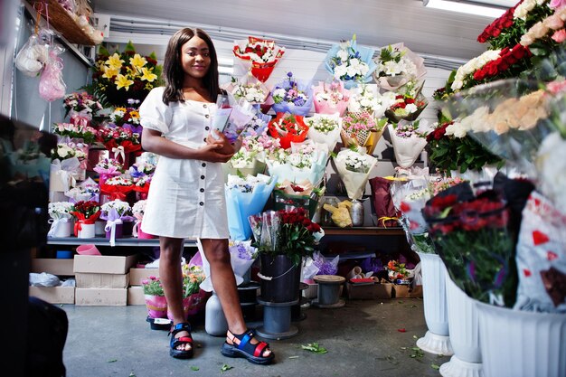 Hermosa chica afroamericana en tierno vestido blanco con ramo de flores en las manos de pie contra el fondo floral en la florería