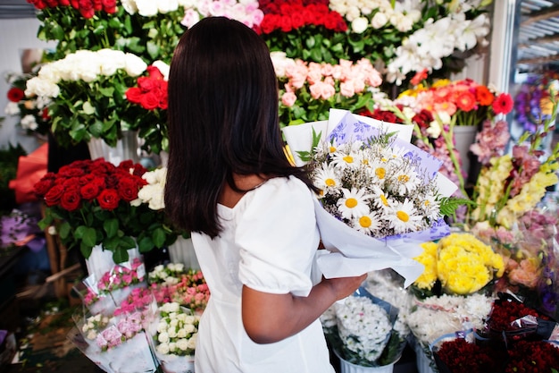 Hermosa chica afroamericana en tierno vestido blanco con ramo de flores en las manos de pie contra el fondo floral en la florería