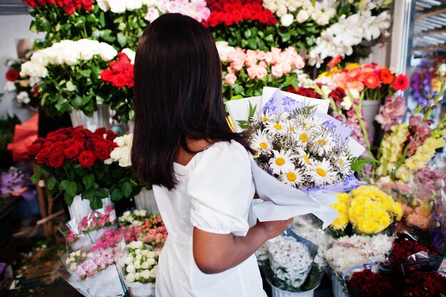 Hermosa chica afroamericana en tierno vestido blanco con ramo de flores en las manos de pie contra el fondo floral en la florería