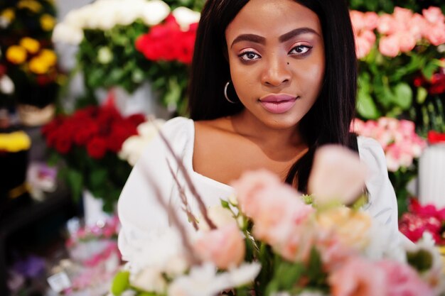 Hermosa chica afroamericana en tierno vestido blanco con ramo de flores en las manos de pie contra el fondo floral en la florería