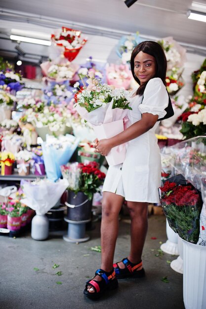 Hermosa chica afroamericana en tierno vestido blanco con ramo de flores en las manos de pie contra el fondo floral en la florería