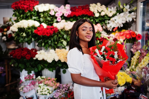Hermosa chica afroamericana en tierno vestido blanco con ramo de flores en las manos de pie contra el fondo floral en la florería